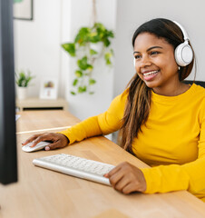 Happy young woman working from home, enjoying music with wireless headphones while using a desktop...