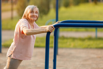 A happy 87-year-old senior woman performing an assisted push-up or leaning exercise on blue outdoor parallel bars, looking at the camera. Healthy and strong retirement.