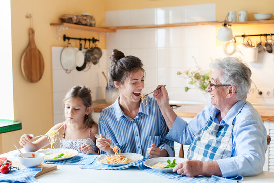 Happy multigenerational family eating pasta together at home kitchen, having fun. Grandmother feeding adult daughter, child enjoying comfort food. Candid, authentic lifestyle, joyful mealtime moment - Powered by Adobe