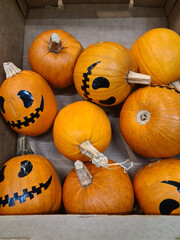 A collection of orange pumpkins with painted faces, arranged in a wooden box. The pumpkins have various expressions, suitable for Halloween decorations.