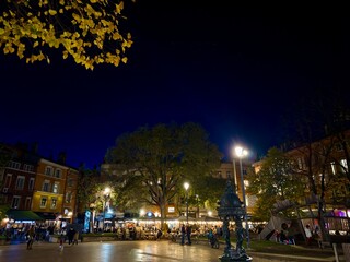 Vibrant Outdoor Night Scene Capturing A Lively Square With People Enjoying Cafes And Restaurants Surrounded By Historic Architecture And Illuminated By Street Lamps