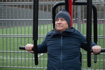 Obraz premium Adult caucasian man performing strength exercise on outdoor machine, staying active and supporting healthy lifestyle and overall fitness. Cold season and rain.