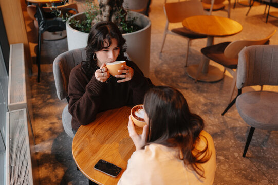 young woman sipping coffee in caf&eacute;, companion listening across round table, soft interior lighting,
