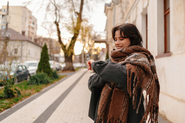 Fototapeta premium smiling woman holding gray cat on sidewalk, wrapped in layered shawl and overcoat, strolling past boutique
