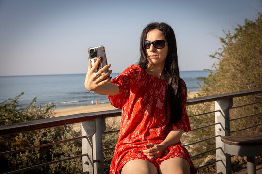 Stylish woman in a red dress taking a selfie with a smartphone by the sea on a sunny day.