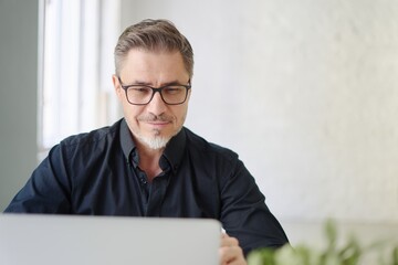 Business portrait - confident businessman sitting at desk in office working with laptop computer. Happy mid adult man in shirt and jacket, smiling.