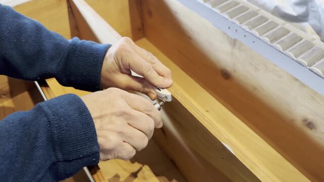 Close-up of hands using a screwdriver to secure a metal hinge onto a wooden furniture piece, demonstrating detailed craftsmanship and DIY construction techniques