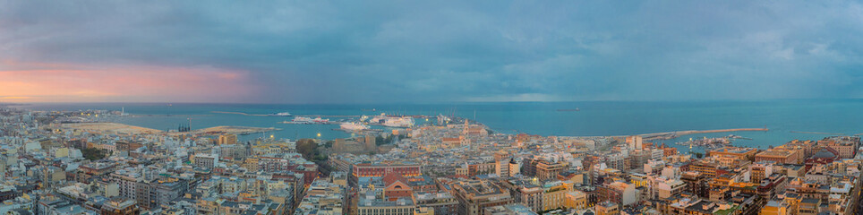 Bari at dusk with a panoramic view of the historic center, bustling port, and calm Adriatic Sea under dramatic clouds and soft sunset light.