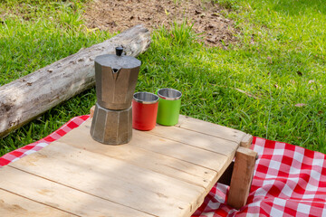 Naklejka premium Outdoor picnic scene with moka pot and metal cups on rustic wooden table over checkered blanket in tropical setting