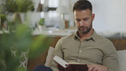 Young man reading book sitting on couch at home.