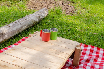 Small rustic picnic setup with metal cups on wooden table over checkered blanket in tropical outdoor setting