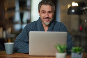 Businessman in home office wearing bathrobe working with laptop computer. Portrait of mature age, middle age, mid adult casual man in 50s, confident happy smiling