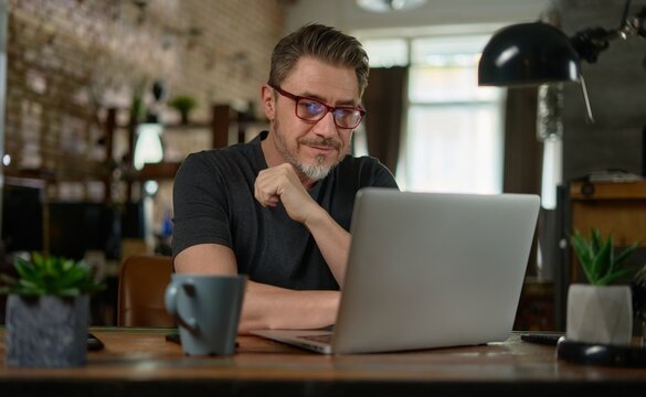 Bearded man working online with laptop computer at home sitting at desk. Home office, browsing internet. Portrait of mature age, middle age, entrepreneur.