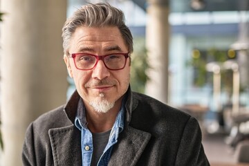 Portrait of happy middle aged man in glasses, standing in office lobby wearing coat.