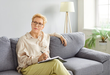 Elderly woman sitting on a sofa writing in a notepad, planning calendar and agenda at home. Focused on planning schedule and weekly plan, she working and engaged in personal tasks.