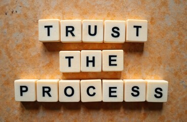 Naklejka premium Scrabble tiles spell out TRUST THE PROCESS on textured orange background. Motivational phrase for business, personal growth, patience, and perseverance.