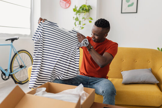 Happy African man opening delivery box with new clothes purchased online at home. Black male customer receiving cardboard parcel box. Online shopping and home delivery concept