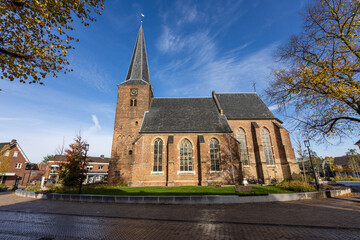 Historic brick church with tall steeple and clock tower, arched windows and slate roof framed by autumn trees, plaza and benches add charm to this serene village scene under clear blue sky.