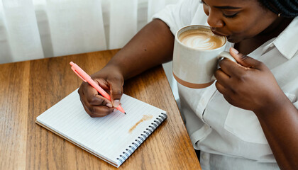 Black woman writing in a journal and drinking a latte from a mug. A person planning their day in a notebook. Morning routine and creative lifestyle concept