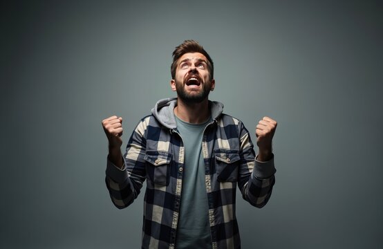 Man looks up screaming with fists clenched in despair and anger. He wears a plaid shirt and hoodie showing extreme frustration and bad luck with a gray background. He gestures with emotion. - Powered by Adobe