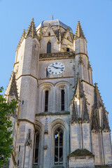 Obraz premium A tall, grey Gothic church Saint Peter in Saintes, France. The building is grey. The photo was taken from the bottom up, the sky is blue The clock on the tower is round and white. With black hands