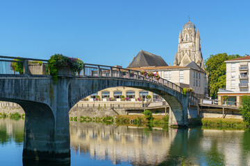 An arched bridge over the Charente River in Saintes, France. The tall Gothic church of Saint Peter is visible in the background There are flower pots hanging on the bridge