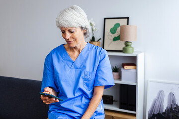 Portrait of senior healthcare worker in blue scrubs relaxing on sofa using mobile phone app or reading an e-mail notification after workday at hospital. Technology and healthcare business concept