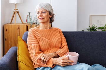 Portrait of mature woman holding coffee mug sitting on sofa in winter in the morning at home