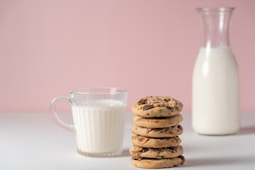 A glass of milk and a stack of cookies sit on a table. The cookies are piled on top of each other, and the milk is in a glass. Jug of milk in the background.
