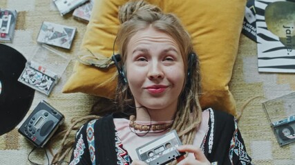 Portrait of cheerful girl in headphones lying on rug, holding cassette tape, enjoying music and smiling at camera surrounded by vintage vinyl records and tapes. Close-up, top view