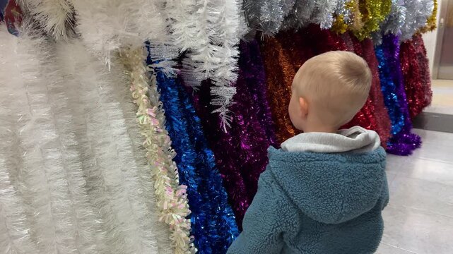 Little curious boy looking and touching multicoloured Christmas tinsel in a shop. Christmas holidays content. 4K