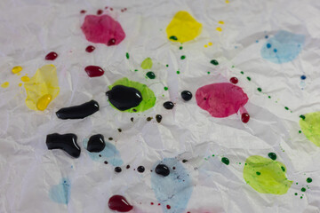 An abstract close-up shot of crumpled white paper or plastic surface, splattered with drops and stains of bright egg dyes – red, yellow, green, and blue, including small dark color splatters