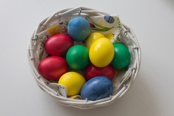 Malý pletený košík, plný sytě obarvených velikonočních vajíček v barvách modré, žluté, zelené a červené. Košík je umístěn na světlém, mírně rozmazaném pozadí