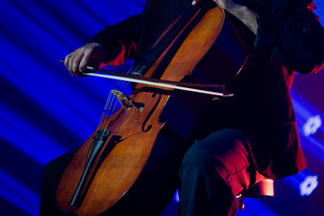 Musician performing on a cello under dramatic blue and red stage lighting with close up focus on the instrument and bow movement in a live concert environment © K