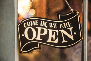Fototapeta premium Closeup of a vintage open sign hanging on a shop window with warm bokeh lights in the background.
