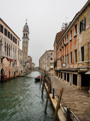 Picturesque view of a Venetian canal, the Rio dei Greci, with the arched bridge and the distinctive leaning bell tower of the Church of San Giorgio dei Greci, standing out against an overcast sky.