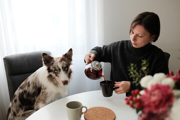 A woman pours tea at a breakfast table while her dog watches attentively, creating a warm domestic moment. The scene reflects companionship, calm routine, and quiet morning atmosphere.