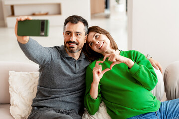 Happy couple taking selfie showing heart symbol sitting on comfortable sofa at home