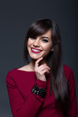 Close-Up Beauty Portrait of a Cheerful Young Brunette Woman Gently Touching Her Chin and Smiling Softly, Wearing Burgundy Lipstick and a Black Beaded Bracelet