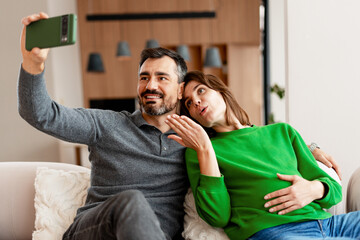 Expecting parents taking selfie at home looking at camera, sitting in living room at home
