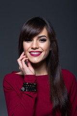 Close-Up Beauty Portrait of a Cheerful Young Brunette Woman Gently Touching Her Chin and Smiling Softly, Wearing Burgundy Lipstick and a Black Beaded Bracelet