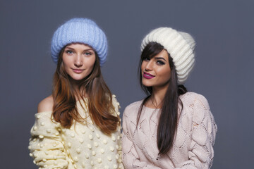 Waist-Up Shot of Two Female Models Displaying Warm Autumn or Winter Knitwear, Featuring a Blue Mohair Hat and Off-Shoulder Yellow Sweater Next to a White Ribbed Beanie and Patterned Beige Pullover