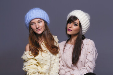 Waist-Up Shot of Two Female Models Displaying Warm Autumn or Winter Knitwear, Featuring a Blue Mohair Hat and Off-Shoulder Yellow Sweater Next to a White Ribbed Beanie and Patterned Beige Pullover
