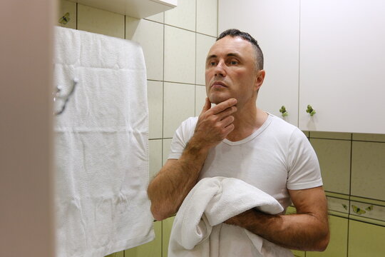 white man shaving with brush and shaving foam looking in the mirror. A man shaves his face in a bathroom with butterflies on the wall and light green tiles.