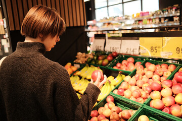 A woman holds an apple while standing at the fruit display in a grocery store. The concept shows everyday shopping and healthy food selection.