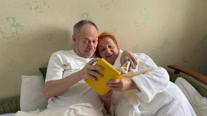 Happy family senior couple relaxing and reading a book together on the bed in bedroom. Elderly husband and wife taking care each other. Retired people healthcare and mental health concept. 4k