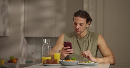 A man sits at a kitchen table, enjoying a colorful breakfast and checking his phone with a relaxed smile