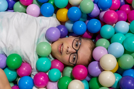 Smiling boy with glasses lying in colorful ball pit surrounded by plastic balls looking at camera at indoor playground. Concept of children entertainment, happy childhood and kids amusement center.