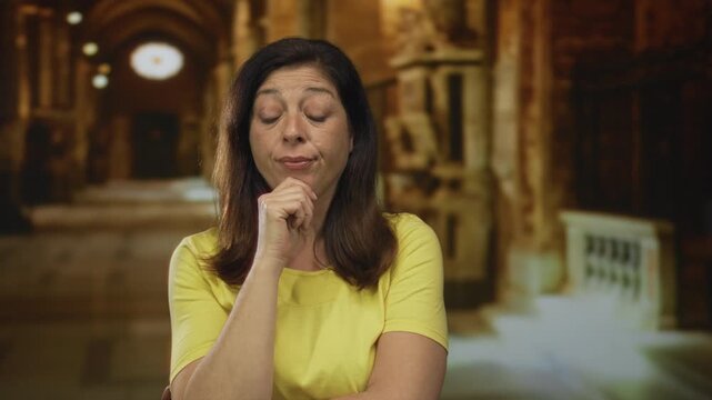 Woman in yellow shirt rests hand on chin in church building illuminated by soft lights; contemplation.