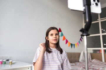child girl recording video blog at home, talking to smartphone camera cozy room with colorful bunting decor. Creative childhood activity, communication, modern lifestyle and digital content creation
